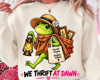 Puede incluir: Una sudadera color crema con el gráfico de una rana verde con sombrero de paja con una margarita y un chal estampado. La rana lleva una linterna encendida y una bolsa con el texto Thrift Finds = Happy Vibes. El texto dice WE THRIFT AT DAWN.