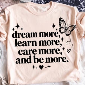 May include: A light peach-colored t-shirt featuring black text that reads, "dream more, learn more, care more, and be more." The design includes a black butterfly graphic, several four-pointed star icons, and a small heart icon near the text.