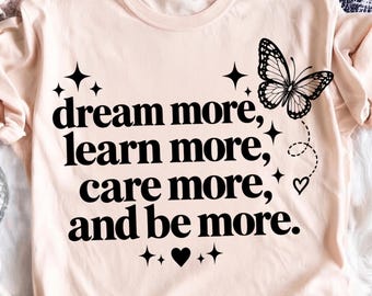 Puede incluir: Una camiseta de color melocotón claro con texto en negro que dice: "dream more, learn more, care more, and be more." El diseño incluye un gráfico de mariposa negra, varios iconos de estrellas de cuatro puntas y un pequeño corazón.