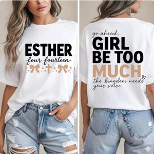 May include: A white short-sleeve t-shirt featuring black and tan text. The front displays ESTHER four fourteen with bow and cross icons. The back reads go ahead, GIRL BE TOO MUCH the kingdom needs your voice in black and tan lettering.