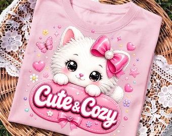 Puede incluir: Una camiseta rosa claro con un gatito blanco de ojos grandes y un lazo rosa en la cabeza. El gatito apoya sus patas sobre un cartel ovalado rosa con el texto "Cute & Cozy" y un pequeño lazo. El diseño incluye corazones, estrellas y flores.