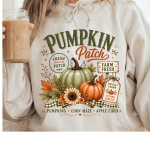 Op de afbeelding: Een crèmekleurige hoodie met een grafische print met de tekst PUMPKIN Patch, FRESH FROM THE PATCH, FARM FRESH en LOCALLY GROWN AUTUMN HARVEST. Het ontwerp bevat een groene pompoen, een oranje pompoen, een zonnebloem en diverse bladeren.