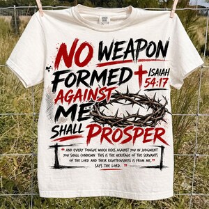 Puede incluir: Una camiseta de manga corta color crema con el texto en negro y rojo NO WEAPON FORMED AGAINST ME SHALL PROSPER, una cruz roja, una corona de espinas marr&oacute;n y la escritura Isaiah 54:17 con texto b&iacute;blico adicional impreso debajo.