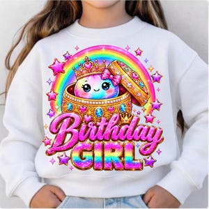 May include: A white long-sleeve sweatshirt featuring a graphic of a cartoon character wearing a crown inside a jewelled gold box, surrounded by a rainbow and stars. The text "Birthday GIRL" is printed in large, pink, glittery, stylised letters.