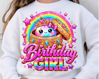 Puede incluir: Una sudadera blanca de manga larga con un gráfico de un personaje de dibujos animados con corona dentro de una caja dorada con joyas, rodeado por un arcoíris y estrellas. El texto "Birthday GIRL" está impreso en letras rosas brillantes.
