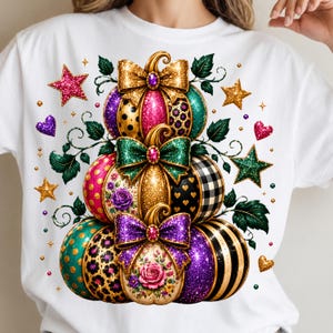 May include: A white t-shirt featuring a graphic of three stacked pumpkins decorated with glitter, bows, and various patterns including leopard print, polka dots, plaid, and floral designs. The image is accented with colorful stars and hearts.