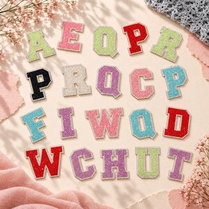 Könnte beinhalten: Eine Sammlung von Buchstaben-Aufnähern im College-Stil mit funkelndem Strass-Finish. Die Buchstaben sind in verschiedenen Farben erhältlich, darunter Rot, Pink, Hellgrün, Hellblau, Lila, Schwarz und Silber, jeweils mit einem weißen Rand.