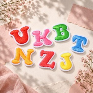 Könnte beinhalten: Eine Sammlung von sieben bestickten Buchstaben-Aufnähern in verschiedenen Farben wie Rot, Pink, Grün, Blau, Orange und Gelb. Jeder Buchstabe weist ein dickes, strukturiertes Stichmuster mit einem weißen Rand auf und ist lose angeordnet.