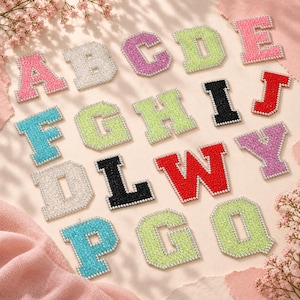 Könnte beinhalten: Eine Sammlung von Buchstaben-Aufnähern im College-Stil, die jeweils mit einem Rand aus kleinen weißen Perlen versehen sind. Die Buchstaben sind in verschiedenen Farben wie Pink, Weiß, Lila, Limettengrün, Türkis, Schwarz und Rot gehalten.