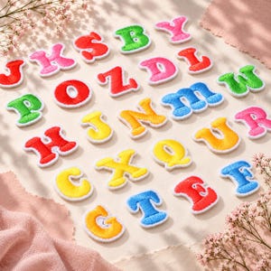 Könnte beinhalten: Eine Sammlung von gestickten Buchstaben-Aufnähern in leuchtenden Farben wie Rot, Pink, Grün, Gelb und Blau. Jeder Buchstabe weist ein dickes, erhabenes Stickmuster mit weißem Rand auf und ist in einer verstreuten Anordnung auf einer hellen Fläche.