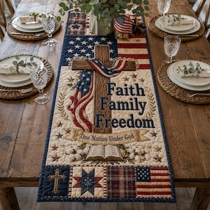 May include: A rectangular quilted table runner featuring a central wooden cross draped with an American flag, the text Faith Family Freedom, and the phrase One Nation Under God. The design includes an open book, cotton bolls, and stars.