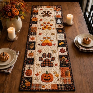 May include: A rectangular quilted table runner featuring a central cream-colored panel with alternating paw prints and pumpkins, surrounded by a border of black, orange, and brown squares decorated with autumn leaves, acorns, ghosts, and turkeys.