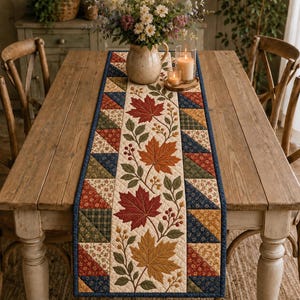 Könnte beinhalten: Ein gesteppter Tischläufer mit einem cremefarbenen Mittelteil, der mit herbstlichen Ahornblättern in Rot-, Orange- und Goldtönen bestickt ist, flankiert von geometrischen Patchwork-Bordüren in Marineblau, Waldgrün und Senfgelb.