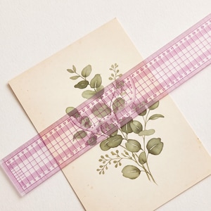 May include: A rectangular off-white card featuring a botanical illustration of green eucalyptus leaves and small berries. A transparent pink plastic ruler with grid markings is placed diagonally across the card, showing a length of 30 centimetres.