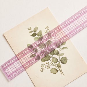 May include: A rectangular off-white card featuring a botanical illustration of green eucalyptus leaves and small berries. A transparent pink plastic ruler with grid markings is placed diagonally across the card, showing a length of 30 centimetres.