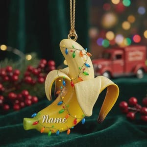Puede incluir: Un adorno navideño con forma de plátano amarillo que cuelga de un cordón dorado. El plátano está parcialmente pelado y envuelto en una cadena de pequeñas luces navideñas multicolores. La palabra Name está impresa en blanco sobre el plátano.