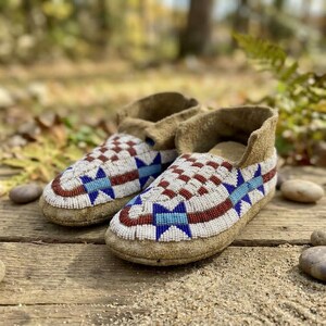 Op de afbeelding: Een paar mocassins van beige suède met ingewikkeld kralenwerk op de wreef. Het kralenwerk toont een geometrisch sterpatroon in witte, donkerblauwe, lichtblauwe en donkerrode glaskralen, die de voorkant van het zachte leren schoeisel bedekken.