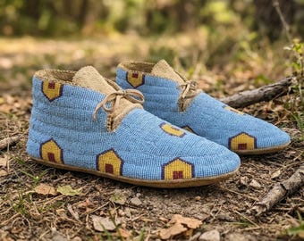 May include: A pair of light blue moccasins featuring a repeating pattern of small yellow and maroon house-shaped motifs. The footwear is crafted with intricate beadwork, tan leather trim, and matching tan leather laces at the front.