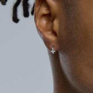 May include: A small silver hoop earring featuring a delicate butterfly-shaped charm adorned with a single clear, sparkling stone at its centre. The earring is secured through the earlobe, showcasing a polished metallic finish and a refined design.