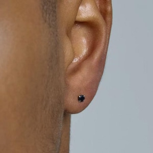 May include: A small, round, dark-colored stud earring featuring a single faceted gemstone set in a metallic prong setting, positioned centrally on the earlobe. The earring has a minimalist design with a polished finish and a secure backing.