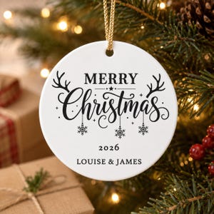 May include: A round white ceramic ornament featuring black text that reads Merry Christmas, the year 2026, and the names Louise & James. The design includes black illustrations of reindeer antlers and hanging snowflakes, suspended by a gold cord.
