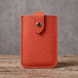 Puede incluir: Un tarjetero rectangular de cuero naranja con textura granulada. El artículo cuenta con un cierre de solapa vertical asegurado por un único botón de presión metálico de tono plateado centrado. Los bordes tienen costuras naranjas.
