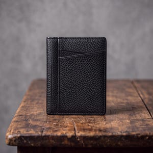 Puede incluir: Un tarjetero de cuero negro con textura granulada. El accesorio rectangular presenta una costura vertical en el lado izquierdo y una ranura diagonal en la parte frontal, diseñado para guardar tarjetas en un perfil delgado y compacto.