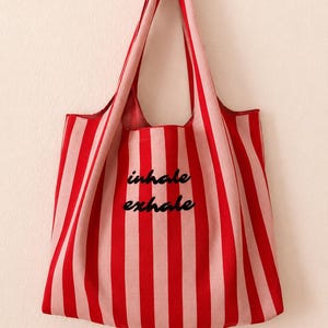 Könnte beinhalten: Eine Tragetasche mit vertikalen Streifen in abwechselnden Rot- und Hellrosatönen. In der Mitte der Tasche befindet sich der Schriftzug "inhale exhale" in einer schwarzen Schreibschrift. Die Tasche hat zwei integrierte Schulterriemen.