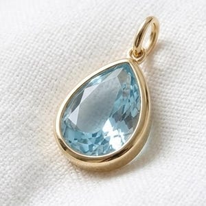 May include: A teardrop-shaped pendant featuring a faceted light blue gemstone set in a polished gold metal frame. The pendant includes a small circular gold bail at the top for attachment to a chain, showcasing a clear, reflective surface.