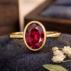 Op de afbeelding: Een gouden ring met een grote, ovaal geslepen, dieprode edelsteen in een gladde zetting. De ringband is van gepolijst goud en de steen heeft meerdere facetten die het licht weerkaatsen, wat zorgt voor een levendige, verzadigde karmozijnrode kleur.