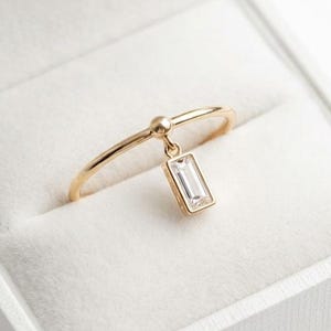 Op de afbeelding: Een dunne gouden ring met een enkele bungelende, heldere baguette-geslepen edelsteen. De ring heeft een delicate band met een klein bolvormig accent waar de rechthoekige steenbedel aan vastzit, rustend in een wit met fluweel bekleed doosje.