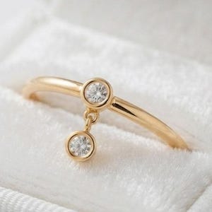 Op de afbeelding: Een gouden ring met een dunne band, een centraal geplaatste ronde heldere steen in een gladde zetting en een kleinere bijpassende ronde heldere steen die aan een kort schakeltje hangt, wat een delicaat hangend ontwerp vormt in een wit fluwelen doosje.