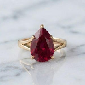 Op de afbeelding: Een gouden ring met een grote, peervormige, dieprode edelsteen in een klauwzetting. De ringband splitst zich in twee afzonderlijke delen naarmate deze de centrale steen nadert, wat zorgt voor een strakke en verfijnde uitstraling van dit sieraad.
