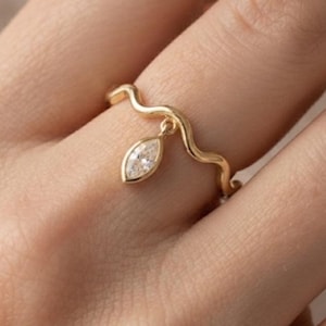 Könnte beinhalten: Ein goldener Ring mit einem dünnen, gewellten Banddesign. Ein einzelner, klarer Edelstein im Marquise-Schliff ist in der Mitte der Welle aufgehängt und hängt vertikal wie ein kleiner Anhänger. Das Metall hat ein poliertes Finish und der Stein ist gefasst.