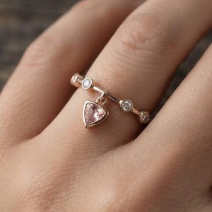 Puede incluir: Un anillo de oro rosa con una banda fina engastada con varias piedras pequeñas, redondas y transparentes. Una sola piedra preciosa triangular de color rosa claro cuelga de la banda como un dije, suspendida por una pequeña anilla de metal.