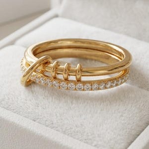 May include: A gold ring featuring a smooth band and a second band encrusted with small clear stones. The two bands are connected by a small gold loop, and the upper band includes three small, vertical gold bars positioned along the top surface.