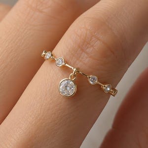 Puede incluir: Un anillo de oro con una banda fina engastada con varias piedras redondas y transparentes pequeñas. Una piedra redonda y transparente más grande cuelga del centro de la banda como un dije, creando un aspecto delicado y refinado.