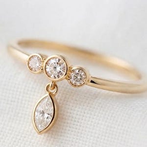 Puede incluir: Un anillo de oro con una banda fina y tres pequeñas piedras redondas engastadas horizontalmente. Una piedra única de talla marquesa cuelga del engaste central, creando un diseño colgante delicado con un acabado de oro amarillo pulido.