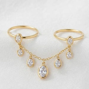 May include: A gold double-finger ring connected by a delicate chain. The piece features seven oval-cut clear stones set in gold bezels, with two stones attached to the ring bands and five dangling from the chain in a cascading arrangement.