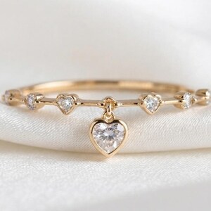 Puede incluir: Un anillo de tono dorado con una banda delgada adornada con pequeñas piedras transparentes en forma de corazón a intervalos. Un dije colgante más grande en forma de corazón con una piedra transparente cuelga del centro de la banda.