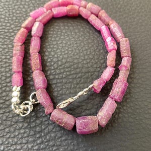 May include: A necklace composed of cylindrical, rough-cut pink ruby beads strung together. The strand features a silver-toned metal lobster claw clasp and a small circular jump ring at the end, providing a secure closure for the jewelry piece.