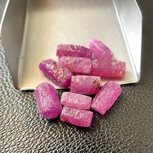 Op de afbeelding: Een verzameling kleine, ruwe, cilindervormige roze robijn-edelstenen met een getextureerd, kristallijn oppervlak. De stenen liggen gegroepeerd, deels in een zilverkleurige metalen schep, en vertonen diverse tinten levendig magenta.