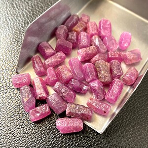 Könnte beinhalten: Eine Ansammlung kleiner, roher, zylindrischer, rosaroter Edelsteine, die in einer Metallschaufel verstreut sind. Die Steine weisen eine raue, texturierte Oberfläche mit unterschiedlichem Grad an Lichtdurchlässigkeit und natürlichen Kristallstrukturen auf.