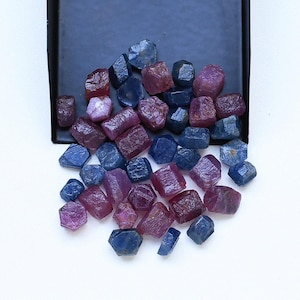 May include: A collection of raw, uncut gemstones in shades of deep blue and vibrant pinkish-red. These small, multifaceted mineral specimens are scattered across a flat surface, with some resting on a dark rectangular tray, displaying natural textures.