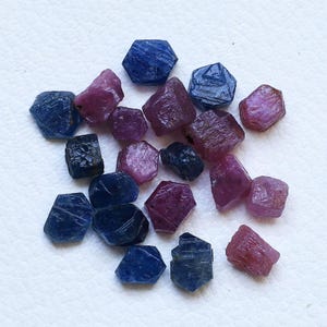 Könnte beinhalten: Eine Ansammlung kleiner, roher, unpolierter Edelsteine in tiefen Blautönen und leuchtendem Pink-Rot. Die Steine weisen unregelmäßige, facettierte Formen mit natürlichen Texturen auf und variieren in Größe sowie in ihrer Lichtdurchlässigkeit.