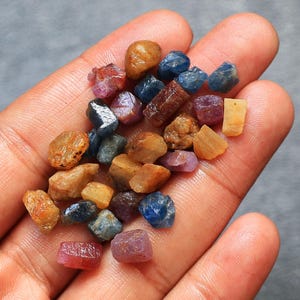 Könnte beinhalten: Eine Sammlung kleiner, roher, ungeschliffener Edelsteine in verschiedenen Farben, darunter tiefes Blau, rötliches Pink, Bernstein und durchscheinendes Gelb. Diese rauen, unregelmäßigen Mineralexemplare weisen natürliche Texturen und vielfältige Formen auf.