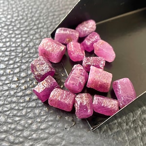 May include: A collection of small, raw, unpolished ruby gemstones with a vibrant pinkish-red hue. The stones are irregular in shape and texture, featuring natural facets and crystalline surfaces, resting inside a dark metal scoop.