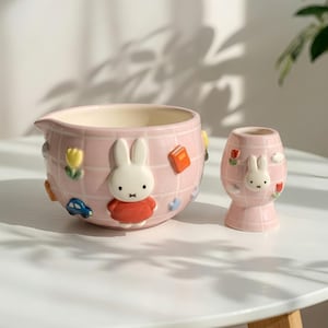 Könnte beinhalten: Eine Keramik-Rührschüssel und ein passender Eierbecher, beide mit einem hellrosa Gittermuster. Die Schüssel zeigt eine erhabene Miffy-Figur in einem roten Kleid, umgeben von kleinen 3D-Symbolen wie einer gelben Tulpe, einem blauen Auto und einem Buch.