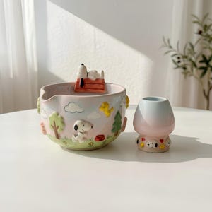 May include: A ceramic bowl featuring a 3D Snoopy on a red doghouse, with embossed trees, clouds, and Woodstock on the exterior. Beside it sits a small, ombre-colored ceramic cup with a base decorated with Snoopy and Woodstock character faces.