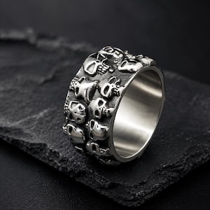 Peut inclure: Une large bague en métal argenté présentant un motif répétitif de crânes en relief sur toute la bande extérieure. La surface de la bague possède une finition sombre et oxydée dans les zones en creux entre les détails des crânes en relief.